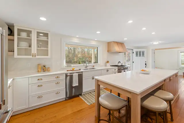 White kitchen by RIKB Design Build with glass-front upper cabinets, a white oak island with upholstered stools and a plaster range hood